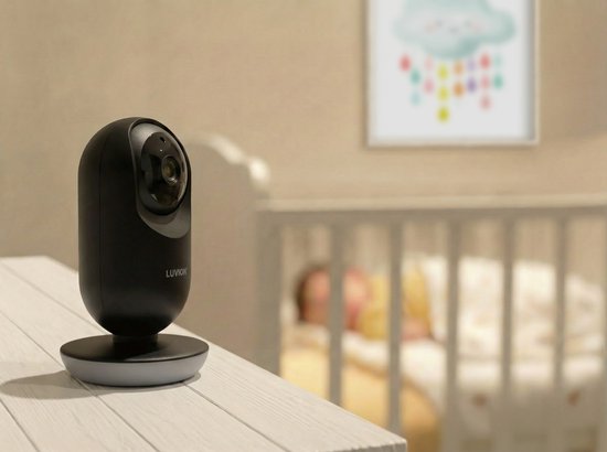 Zwarte Luvion babycamera op een houten tafel, met een baby in een wieg op de achtergrond.