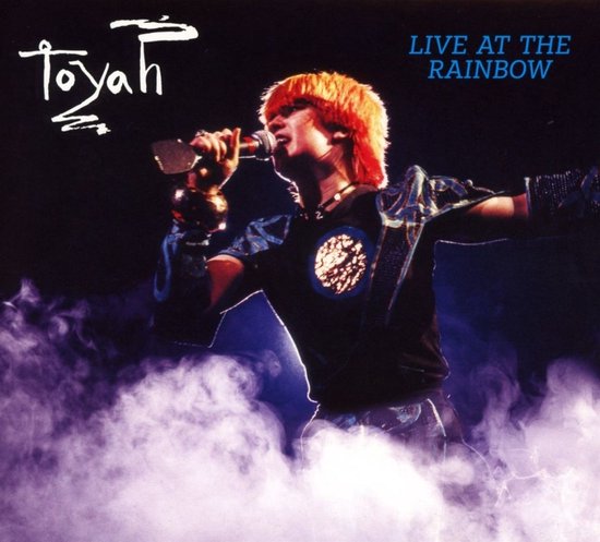 Toyah zingt live in het Rainbow theater, met fel oranje haar, in een zwart shirt met blauwe mouwen en rook op het podium.