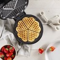 Open zwart wafelijzer met 5 goudbruine hartvormige wafels. Een schaal met aardbeien staat ernaast.