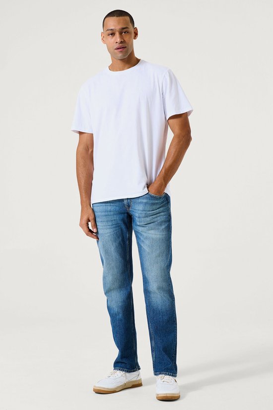 Man met hand in zak, draagt een witte T-shirt en een blauwe jeans met stonewash.