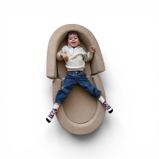 Jongen in beige bank/campingbedje, draagt spijkerbroek en wit vest, lachend.