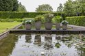 Vijf taupe bloempotten met ribbelpatroon, gevuld met planten en bloemen, staan op een houten vlonder aan een vijver.
