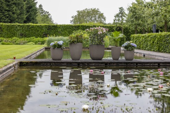 Vijf taupe bloempotten met ribbelpatroon, gevuld met planten en bloemen, staan op een houten vlonder aan een vijver.