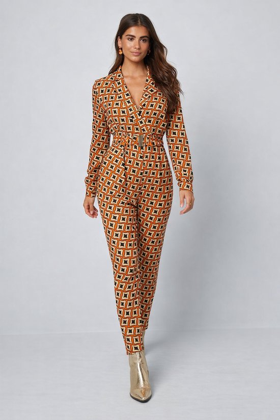 Dames jumpsuit met geometrische print in oranje, beige en zwart. De jumpsuit heeft lange mouwen, een riem en een V-hals. Model draagt gouden laarzen.