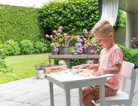 Jongen met blond haar in een oranje shirt, speelt Mikado buiten aan een tafel.