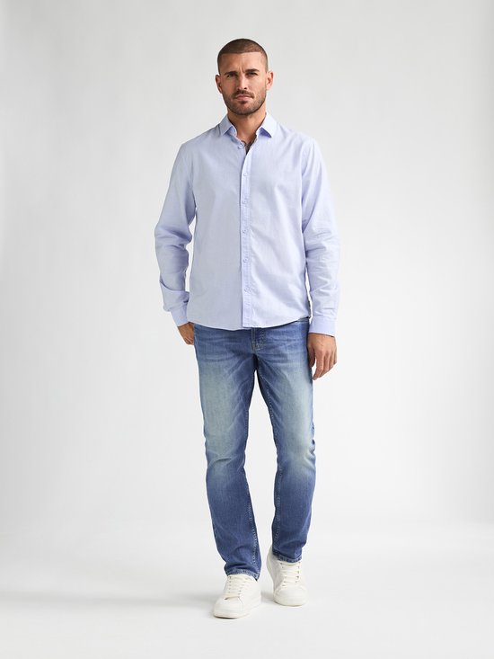 Man in het midden van een lichtblauwe gestreepte blouse en een lichtblauwe spijkerbroek, met witte sneakers. Bekeken vanaf de voorkant.