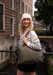 Vrolijke vrouw met krullen en muts, draagt een olijfgroene nylon tas met bruine handvatten.
