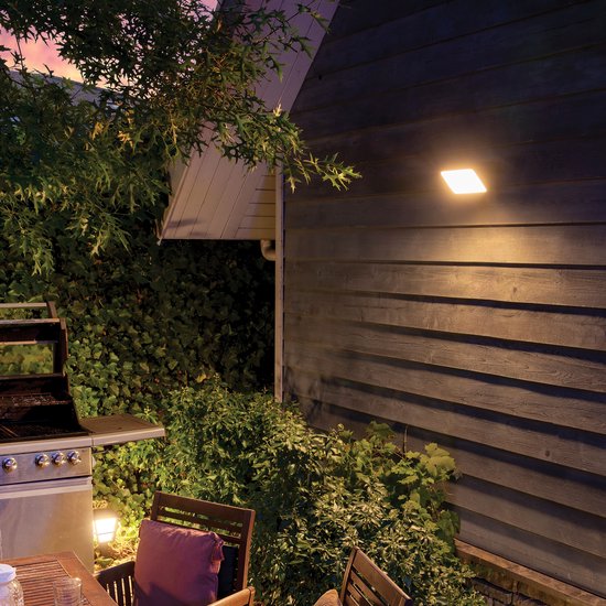 Philips Hue buitenlamp op een houten gevel, naast een eettafel en grill in een tuin.
