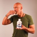 Een gespierde, kalende man met een groene t-shirt neemt een vitamine slice. Hij houdt een fles 'MAN MULTIVITAMIN' vast.