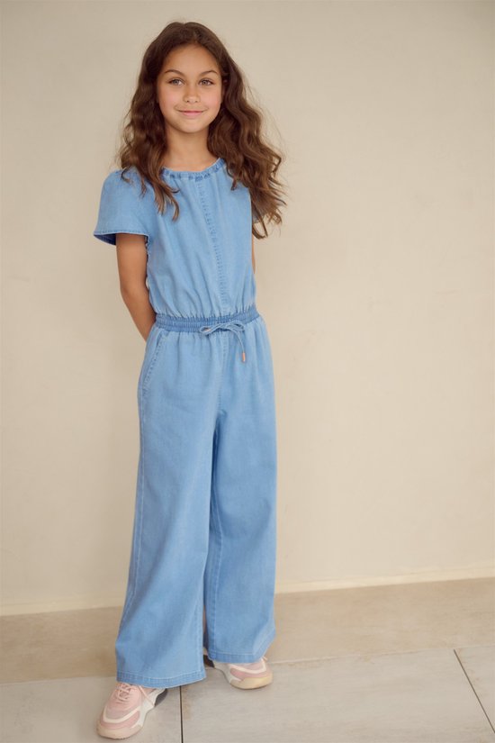 Meisje met lang bruin haar draagt een lichtblauwe denim jumpsuit met korte mouwen en wijde pijpen. Ze draagt roze sneakers.