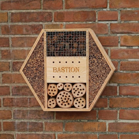 Hexagonaal houten insectenhotel met 'BASTION' gegraveerd, tegen een rode bakstenen muur.