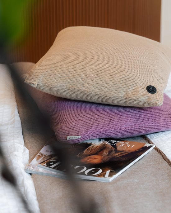 Twee kussens, oudroze en beige, op elkaar gestapeld op een bed. Een tijdschrift ligt ervoor.