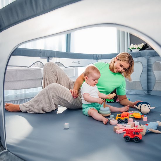 Een vrouw speelt met een baby in een grijze box, omringd door speelgoed.