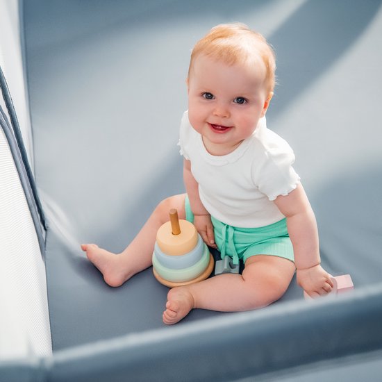 Lachende baby in een grijze box met een gestapeld speelgoed.