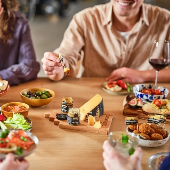 Een man met een glas rode wijn, terwijl hij een stuk kaas prikt. Diverse kazen en hapjes op tafel.