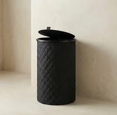 Zwarte, ronde wasmand met ruitpatroon en deksel, tegen een lichte muur.