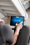 Een vrouw wijst naar een tablet waarop een roei-app met een virtueel landschap wordt weergegeven, zittend op een roeimachine.