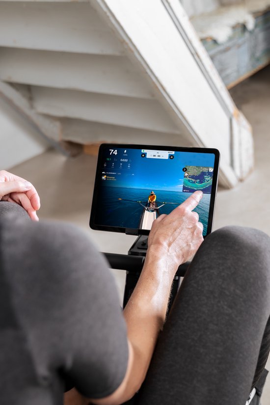 Een vrouw wijst naar een tablet waarop een roei-app met een virtueel landschap wordt weergegeven, zittend op een roeimachine.