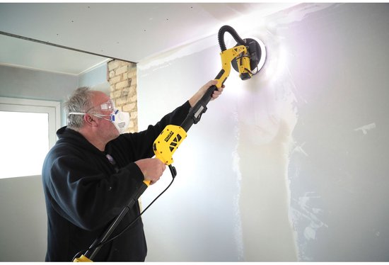 Een man met grijze handschoenen en een zwarte trui schuurt een muur met een gele en zwarte telescoopschuurmachine.