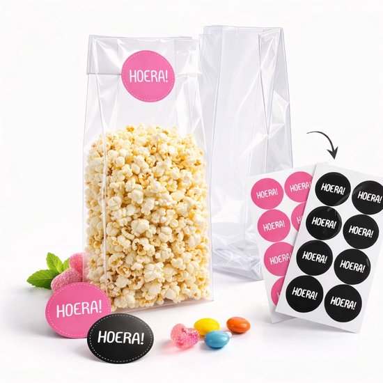 Een transparant blokbodem traktatiezakje gevuld met popcorn en een roze 'HOERA!' sticker, naast een leeg zakje en snoepjes.