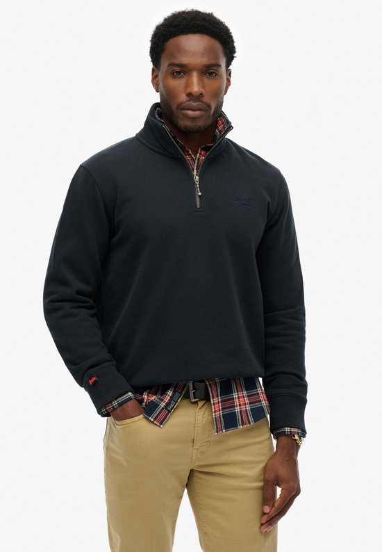 Man met donkerblauwe half-zip trui, geruit hemd, beige broek, riem en instappers.