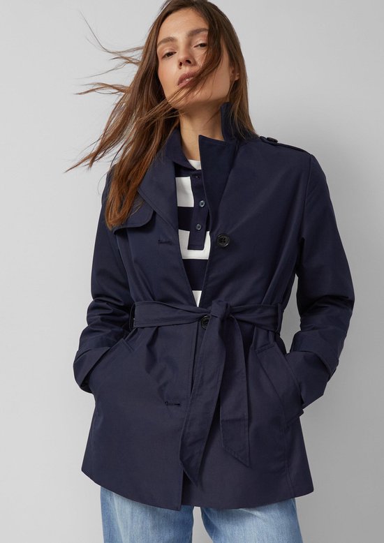Donkerblauwe trenchcoat met ceintuur in de taille, gedragen door een model met wapperend haar.