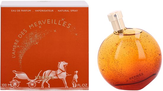 Oranje parfumfles met gouden dop, met sprankelende gouden deeltjes en L'Ambre des Merveilles tekst. Naast een oranje doosje met karos en paard.