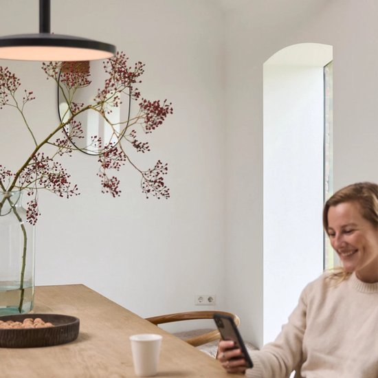 Vrouw in lichte trui lacht naar haar telefoon aan een houten tafel met een vaas met takken en rode bessen.