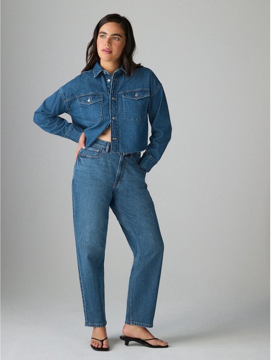 Model staat en draagt een blauwe denim blouse en mom jeans.