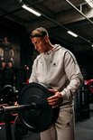 Man met beige trainingsjas en broek, houdt een halterplaat vast in de sportschool.