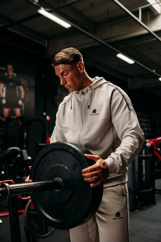Man met beige trainingsjas en broek, houdt een halterplaat vast in de sportschool.