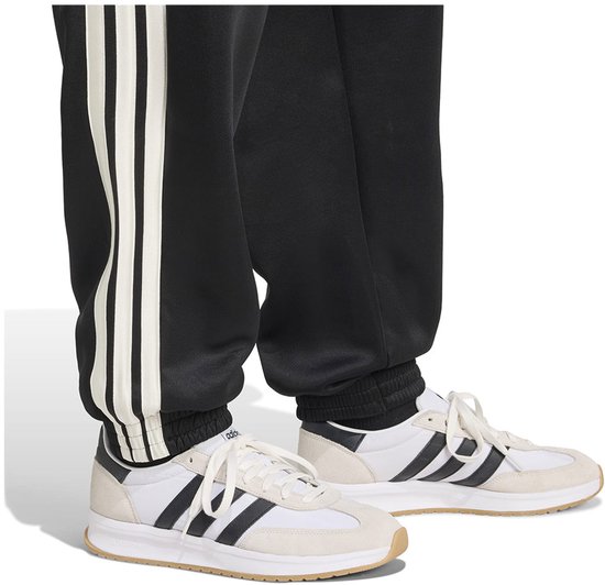 Detail van de broekspijpen van een zwarte trainingsbroek met witte strepen, gedragen met witte sneakers.