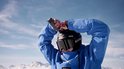 Skiër in blauwe jas, met zwarte helm en ski-bril, past camera op helm aan.