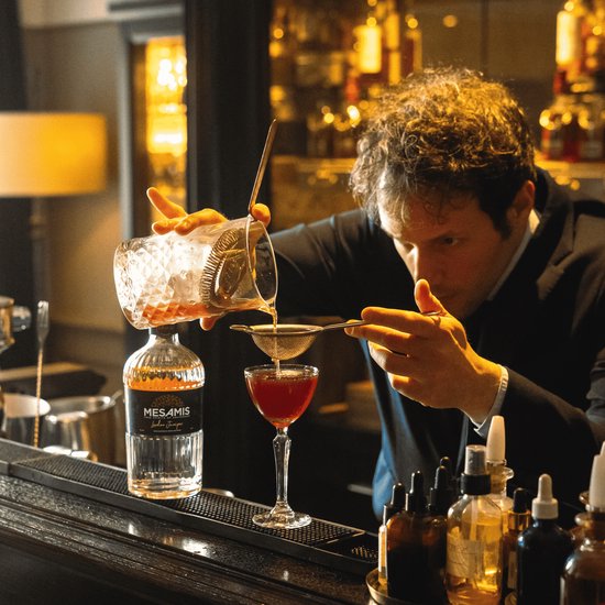 Barman in pak schenkt een rode cocktail in een glas, met een fles Mesamis London Juniper ernaast.