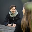 Een man zit aan een tafel met een donkere spijkerjas en een lichtgrijze colsjaal met capuchon, die hij aantrekt.