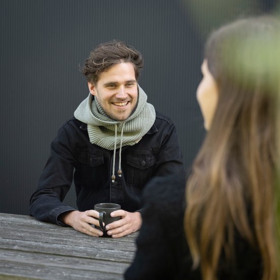 Een man zit aan een tafel met een donkere spijkerjas en een lichtgrijze colsjaal met capuchon, die hij aantrekt.