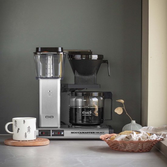 Zilverkleurige Moccamaster Select koffiezetapparaat met glazen kan, naast een mok en een mand met broodjes.