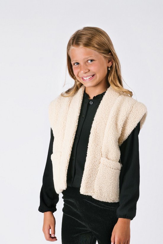 Meisje met blond haar draagt een crème teddy gilet over een zwart shirt. Gilet heeft steekzakken.