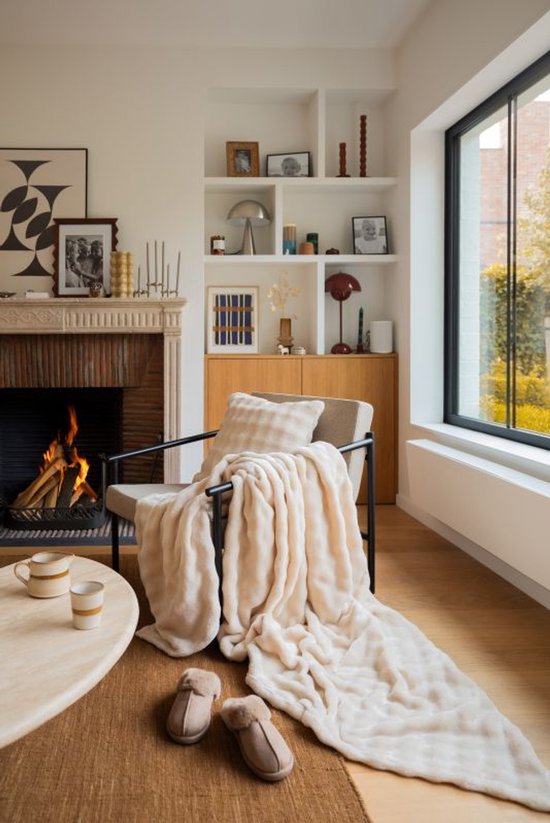Een zacht, crème plaid met golvende textuur ligt over een stoel bij een open haard.