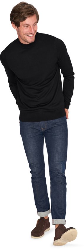 Man lacht, handen achter de rug, draagt zwarte coltrui en jeans.
