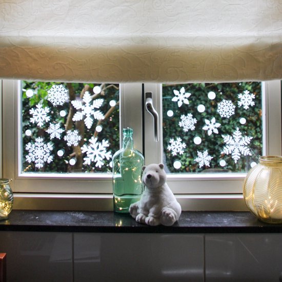 Een raam versierd met witte sneeuwvlokstickers en ronde stickers, met op de vensterbank een berenknuffel, een fles en decoratieve lampjes.