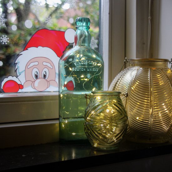 Een groene fles en twee gouden vazen gevuld met warm witte lichtsnoeren, naast een raam met een kerstmansticker.