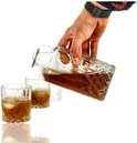 Hand schenkt amberkleurige drank uit een diamantpatroon karaf in twee glazen met ijs. Twee stapelglazen op achtergrond.
