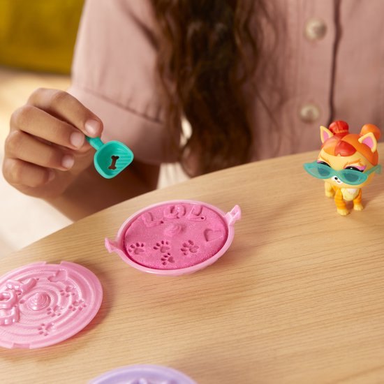 Kind gebruikt een schepje om roze zand te scheppen uit een roze bakje. Oranje kat met zonnebril staat ernaast.