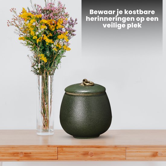 Donkergroene urn met gouden handvat op een houten meubel, naast een vaas met bloemen.