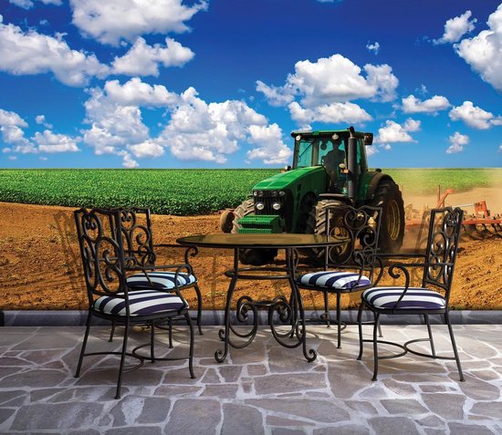 Un tracteur vert John Deere dans un champ labouré avec un champ et un ciel bleu avec des nuages, avec un ensemble de meubles de jardin au premier plan.