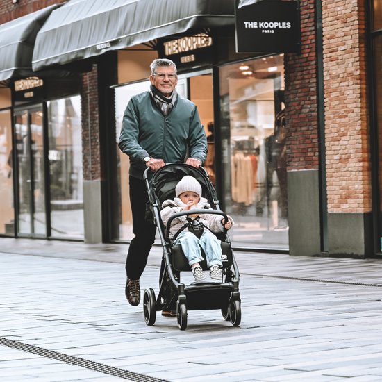 Man met sjaal duwt een zwarte Deryan buggy met een baby erin, op een winkelstraat.