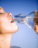 Close-up van een vrouw die water drinkt uit een glazen fles tegen een blauwe hemel.