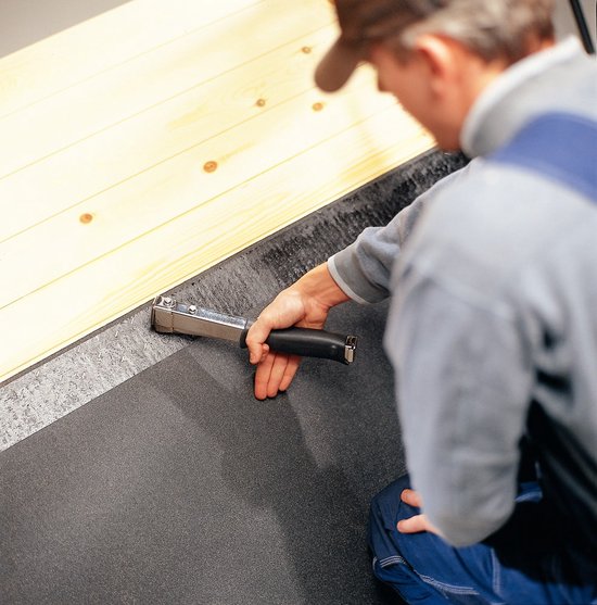 Man met pet en overall bevestigt isolatie met een zilveren hamertacker met zwarte handgreep.