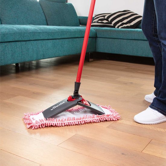 Vileda mop avec manche rouge, microfibre rose et blanche, balayant la poussière d'un plancher en bois, assis sur un canapé.